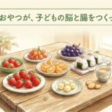 木製のテーブルの上に、カラフルな旬の果物(いちご、みかん、ぶどうなど)と、素朴なおやつ(おにぎり、チーズ)が並んでいる